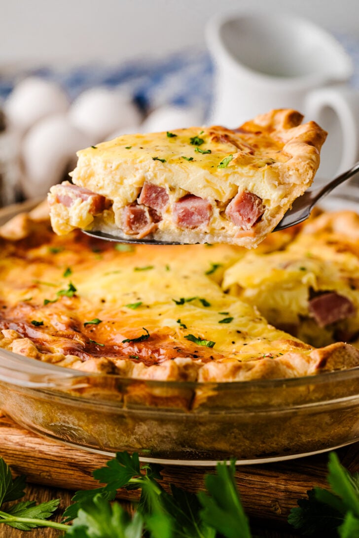 पाई डिश से हैम और पनीर का एक टुकड़ा एक सर्विंग स्पैटुला पर उठाया गया, जिसमें अंदर की मलाईदार फिलिंग और पिघला हुआ पनीर दिखाई दे रहा है।