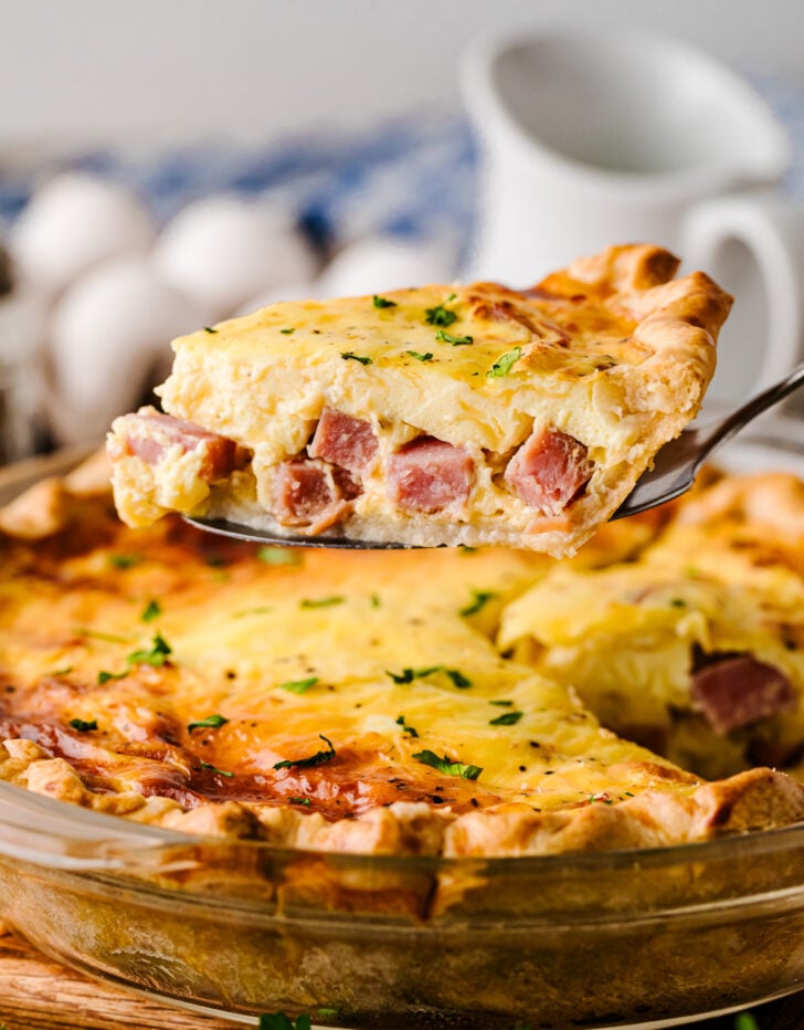 Ham and Cheese Quiche slice on server in glass pie dish, garnished with chives, eggs and pitcher blurred in background