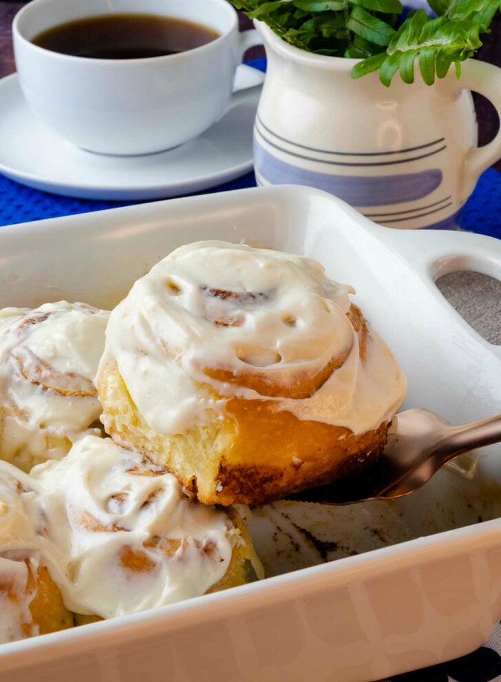 Easy to make Cinnamon Rolls in a pan being scooped with a copper utensil served with a cup of coffee.