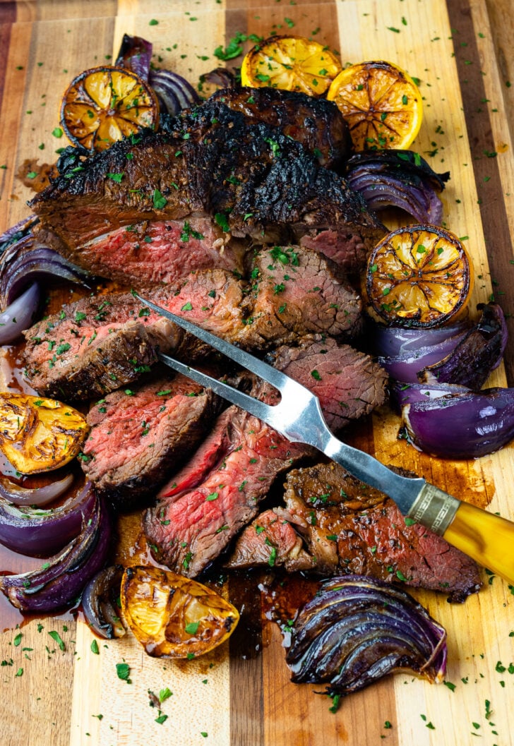 Marinated London Broil with lemons and onions with a serving fork on a wooden board.