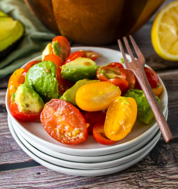 4 white plates stacked with Avocado Tomato Salad and a wooden fork.