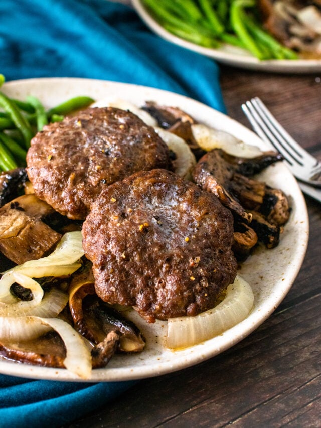 2 hamburger steak patties served with cooked onions and mushrooms and green beans on a plate.