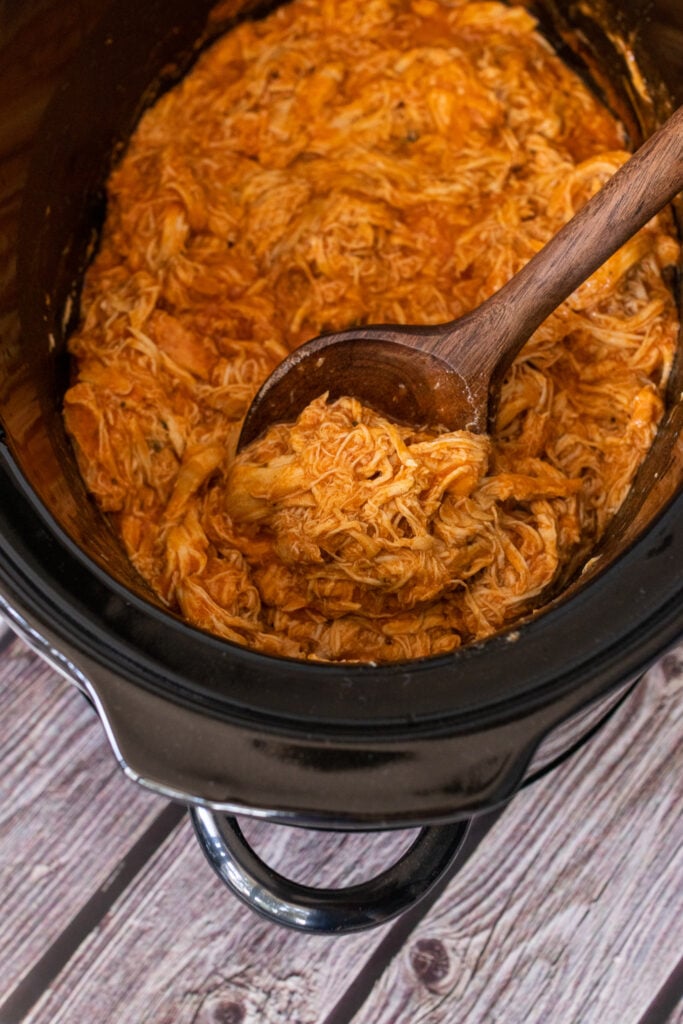 Crock pot buffalo chicken being served with a wooden spoon out of a black crock pot.