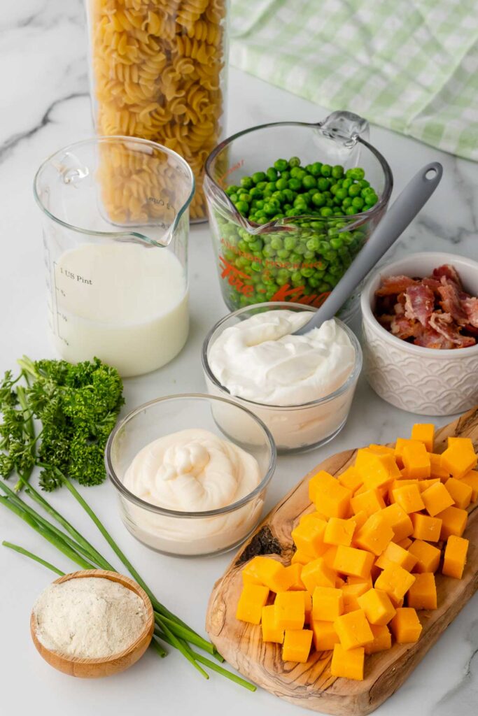Separate white, clear, and wooden bowls are filled with each ingredient; peas, bacon, cheddar cheese, ranch seasoning and few other tasty ingredients. 