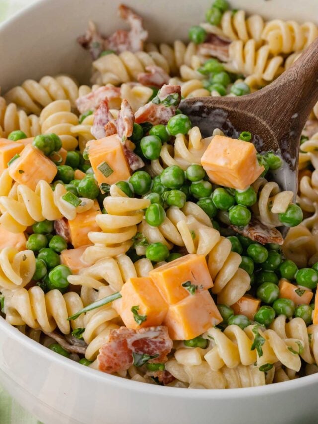 Cropped bacon ranch pasta salad with rotini, peas, cheddar cheese, and bacon bits in white bowl with wooden spoon