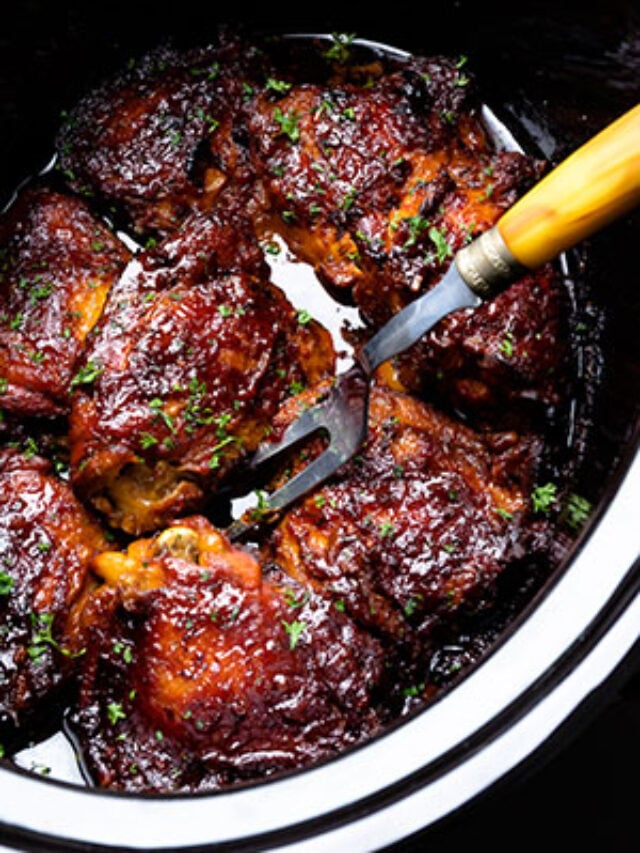 Cropped crock pot chicken thighs in pot with fork, glazed in dark sauce, garnished with green herbs, overhead view