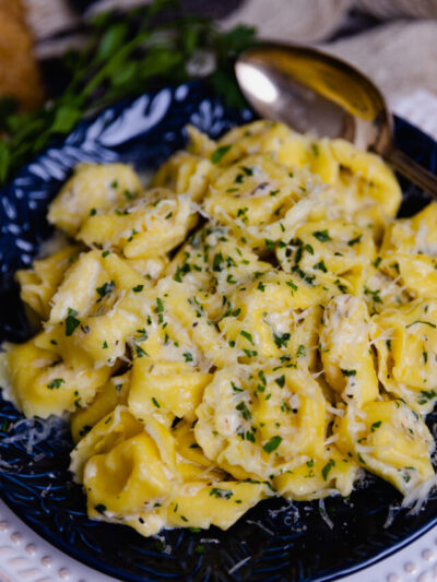 Creamy Cheesy Tortellini made on the stovetop is full of garlic, and herbs with a macaroni and cheese vibe making it a wonderful side dish! Served on a blue plate with a spoon for dinner.