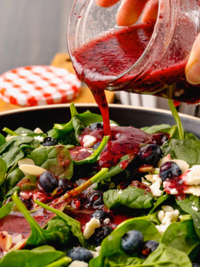 Jar of homemade blueberry vinaigrette salad dressing poured over fresh spinach, sliced almonds and feta cheese for a delicious, refreshing salad.