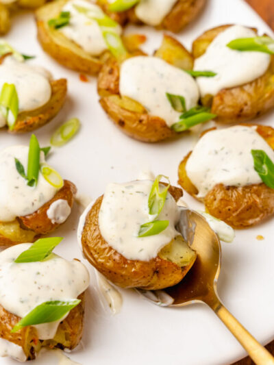 Crushed baby potatoes topped with homemade ranch dressing and chopped green onions served on a white platter and a gold spoon.