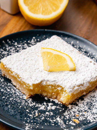 Lemon square topped with powdered sugar and a lemon slice on a black plate.