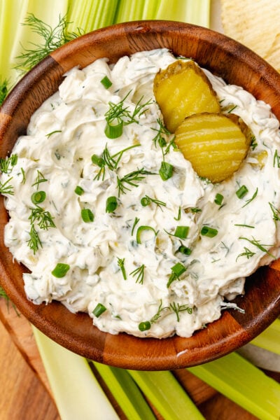 Homemade dill pickle dip served in a wooden bowl with fresh dill and chives garnish along with celery sticks and potato chips.