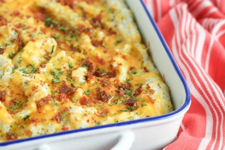 Baked potato casserole close-up in white dish with blue rim, topped with melted cheese, bacon bits, and fresh chives on red striped towel