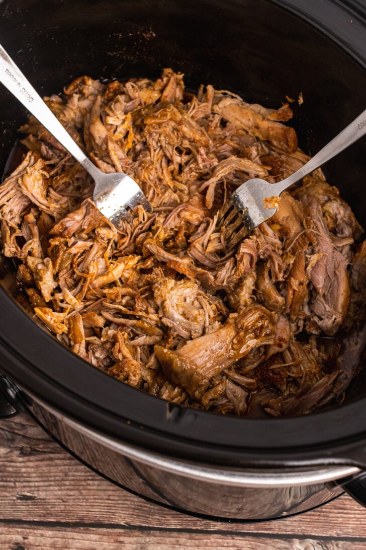 Crock Pot with cooked pulled pork that has been shredded with two forks.