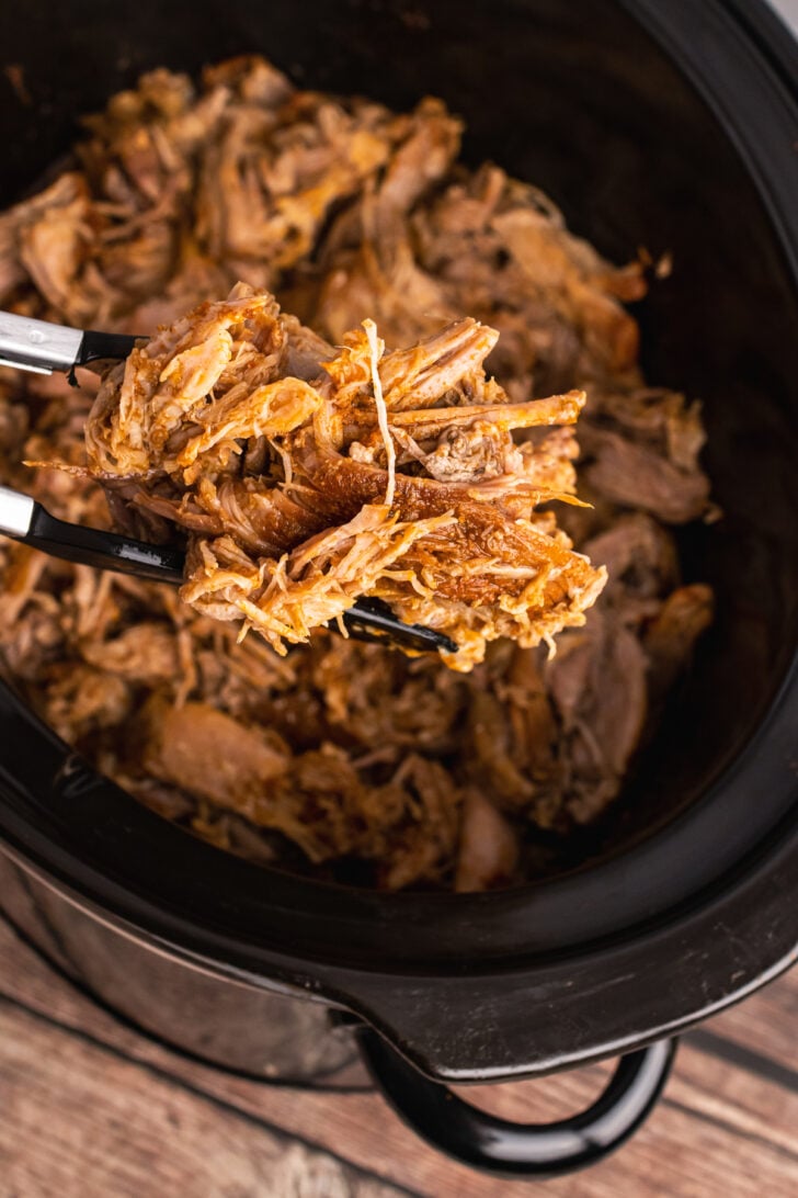 Crock pot with cooked pulled pork that has been shredded, served with tongs.