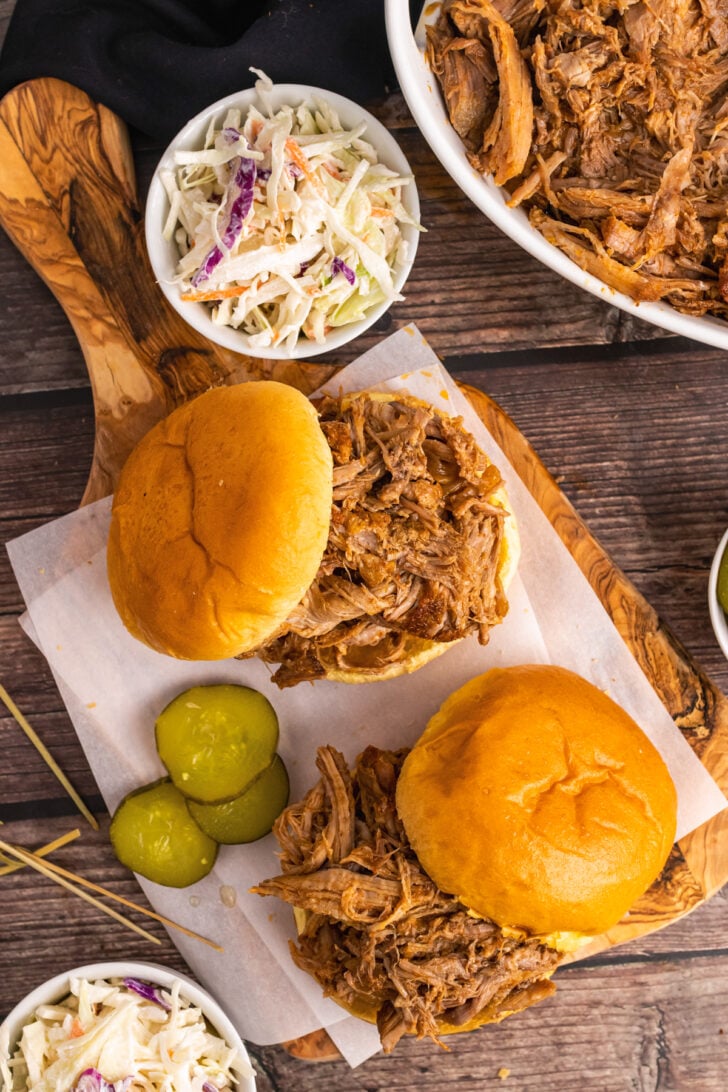 Pulled Pork piled on a round bun on a wooden cutting board served with sliced pickles and coleslaw.