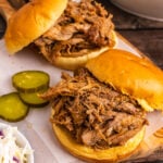 Pulled Pork piled on a round bun on a wooden cutting board served with sliced pickles and coleslaw.
