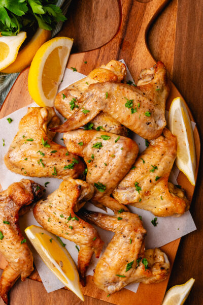 Lemon garlic oven baked chicken wings on a wooden board with lemon slices and topped with chopped parsley.