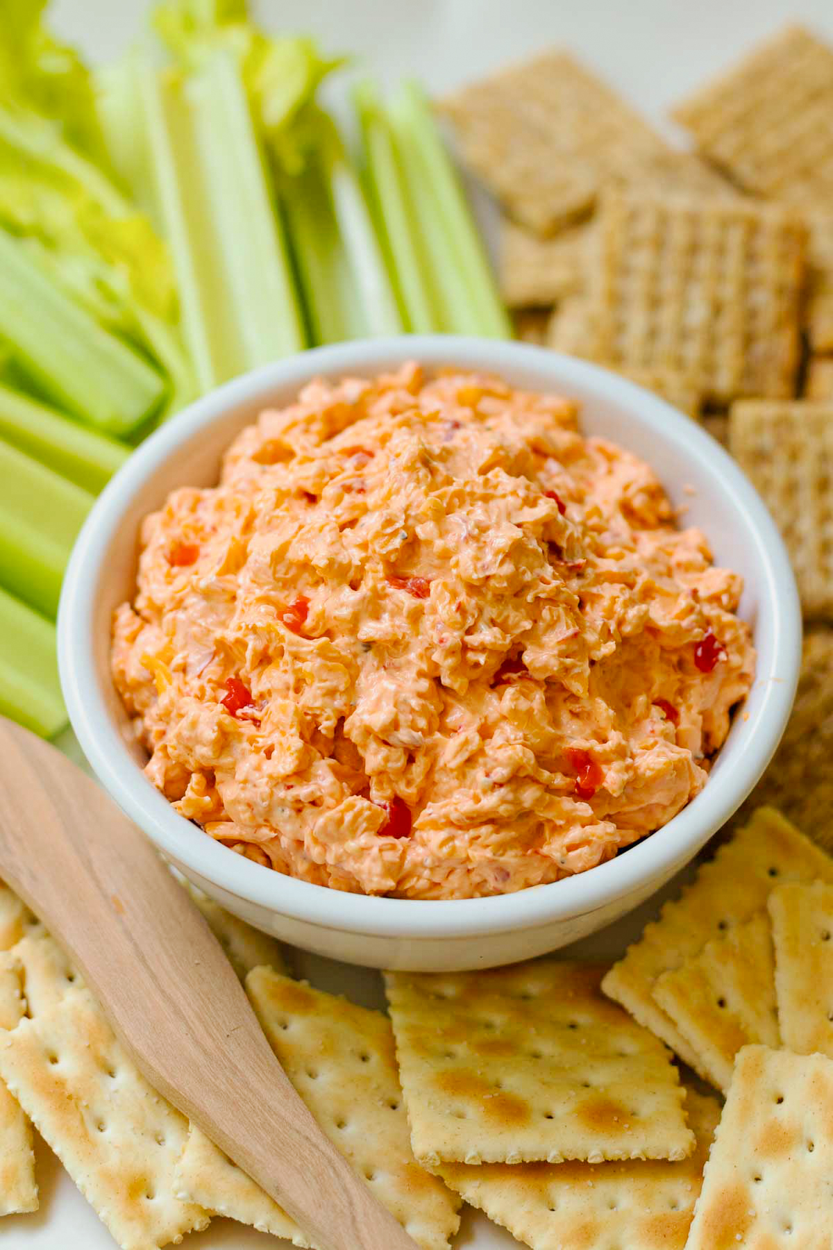 Homemade Pimento Cheese in a white bowl served and wooden knife with saltine crackers and celery sticks on a white platter.