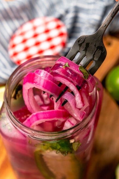Pickled red onions on a fork pulled out of a far that have fresh parsley and jalapeno peppers in the jar.