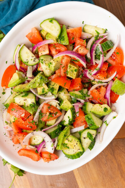 Large white serving bowl with sliced red onions, cucumbers, chopped tomatoes and avocados with homemade dressing and chopped parsley.