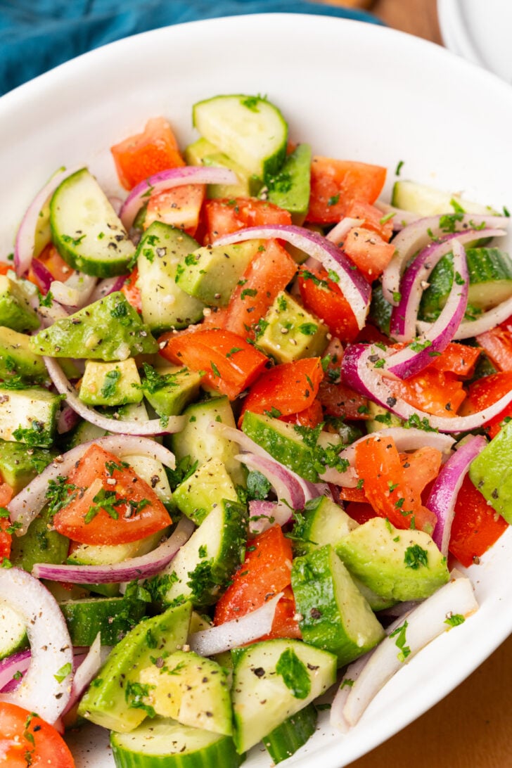 कटा हुआ लाल प्याज, खीरे, कटे हुए टमाटर और एवोकैडो के साथ घर का बना ड्रेसिंग और कटा हुआ अजमोद के साथ बड़ा सफेद सर्विंग बाउल।