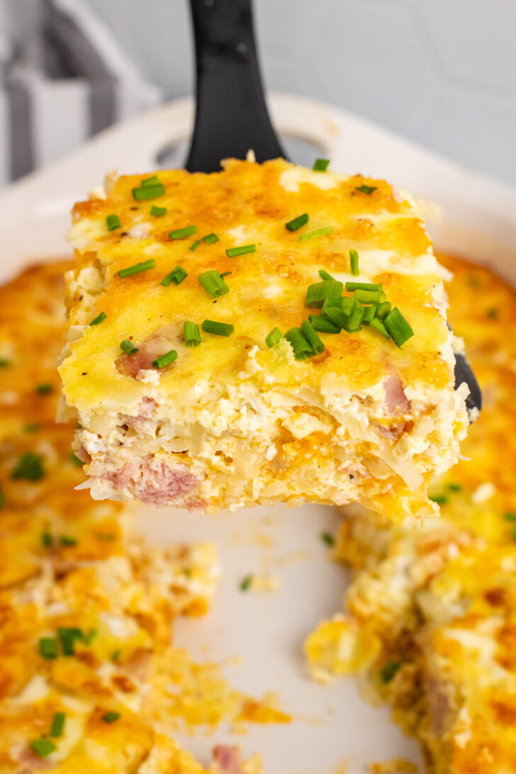 Square slice of ham and potato hashbrown casserole on a black spatula but from a white casserole dish.