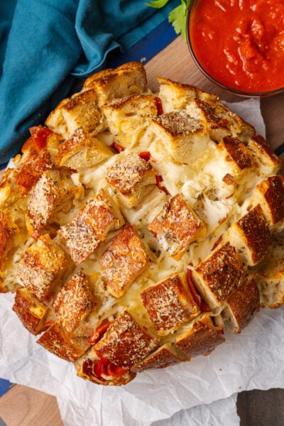 Round load of pepperoni pizza cheese bread with cut squares on parchment paper and a glass bowl of marinara sauce.