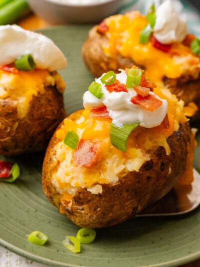 Stuffed baked potatoes with bacon, and cheese then topped with more bacon bits, sour cream and green onion slices on a green plate.