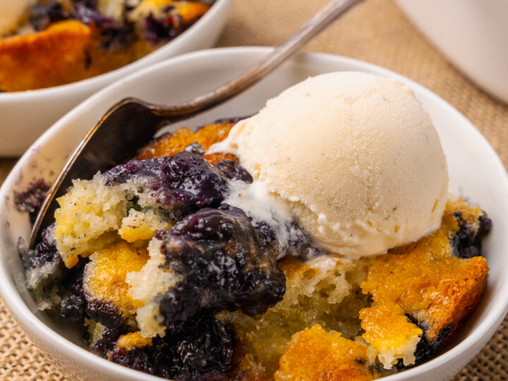 White bowl with blueberry cobbler and a scoop of vanilla ice cream resting on a tan placemat with silver forks.