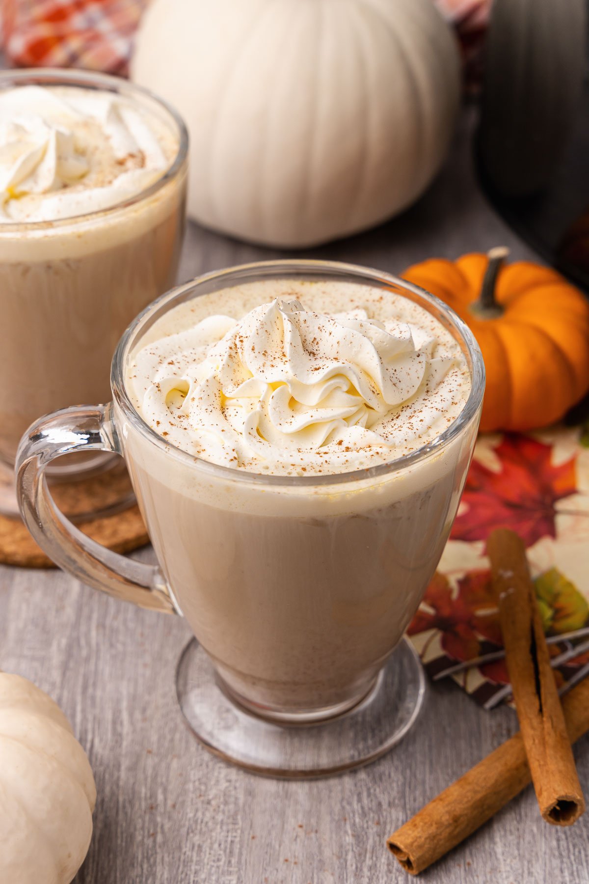 Creamy pumpkin spice latte topped with whipped cream in glass mugs on a wood table with cinnamon sticks.