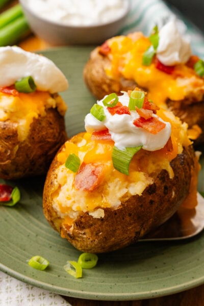Stuffed baked potatoes with bacon, and cheese then topped with more bacon bits, sour cream and green onion slices on a green plate.