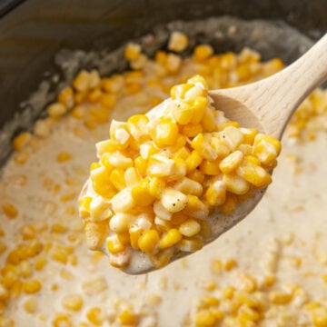 Wooden spoon serving crock pot creamed corn from a black crock pot.