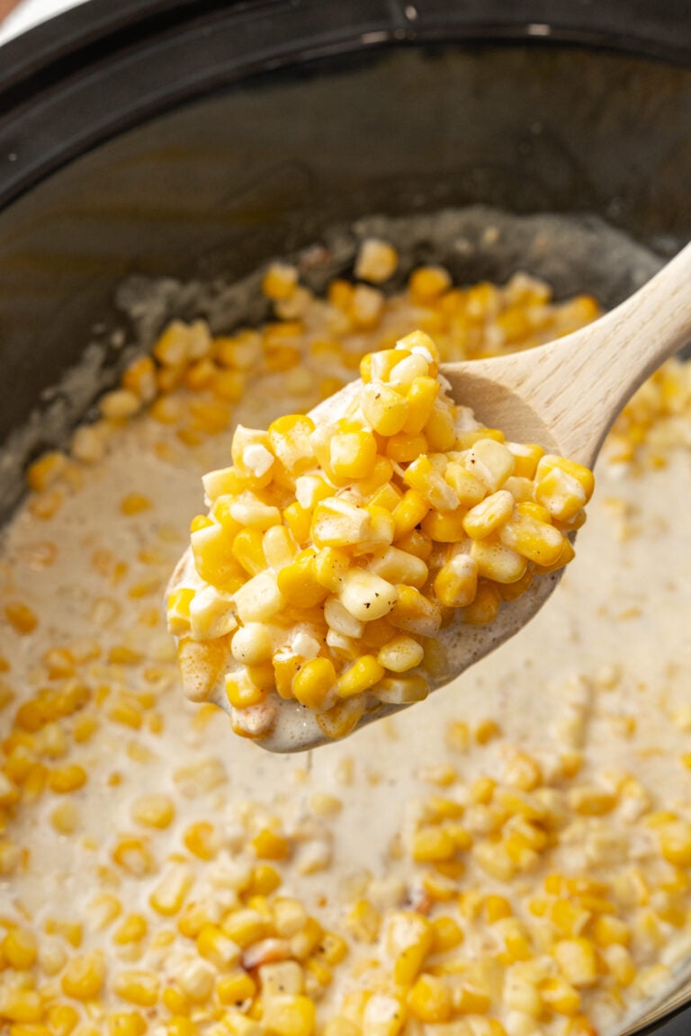Wooden spoon serving crock pot creamed corn from a black crock pot.