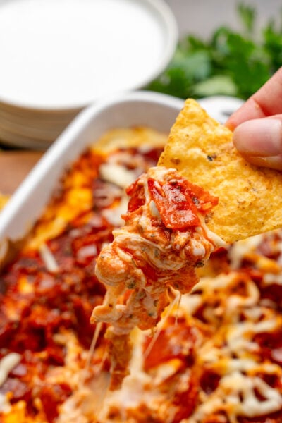 Hand holding a tortilla chip with a scoop of Pepperonit dip with tomato sauce and cheese.