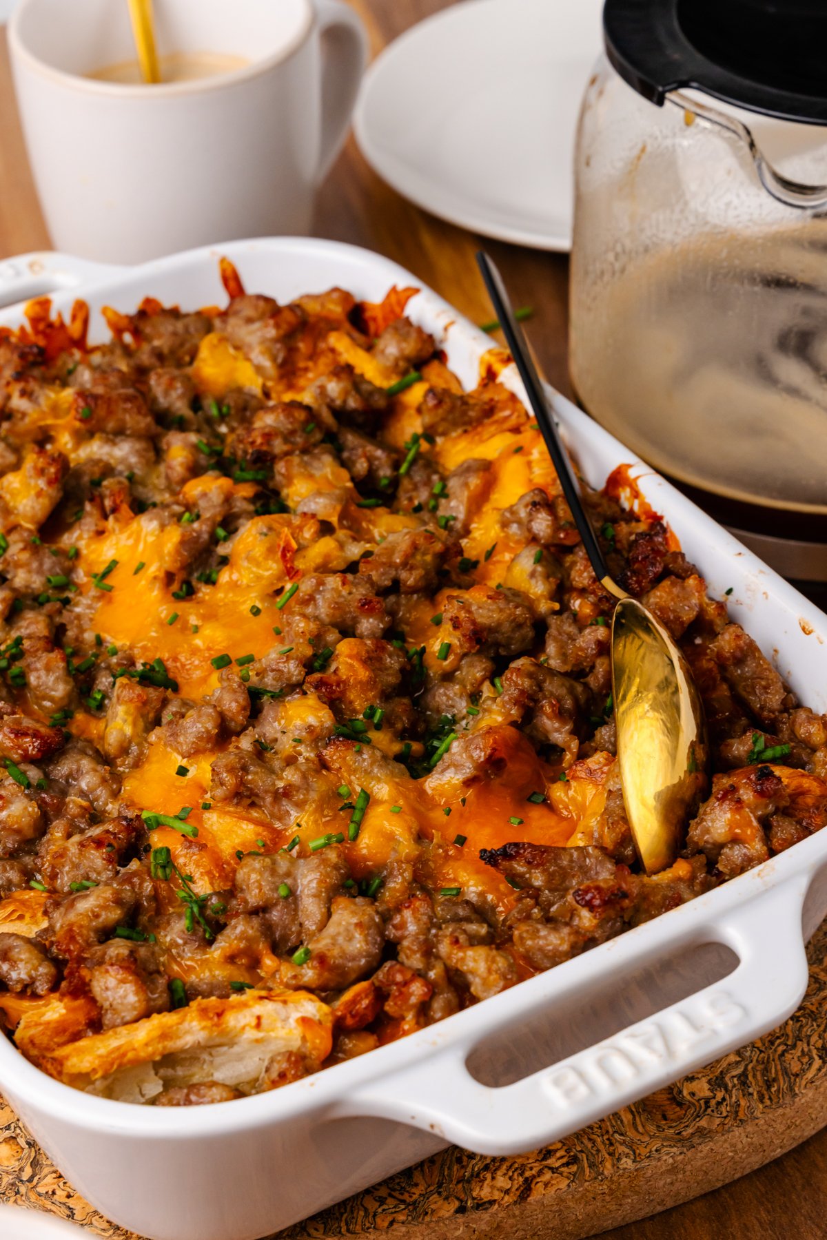 White cast iron baking dish with sausage and cheese croissant casserole topped with chives and served with a gold spoon.