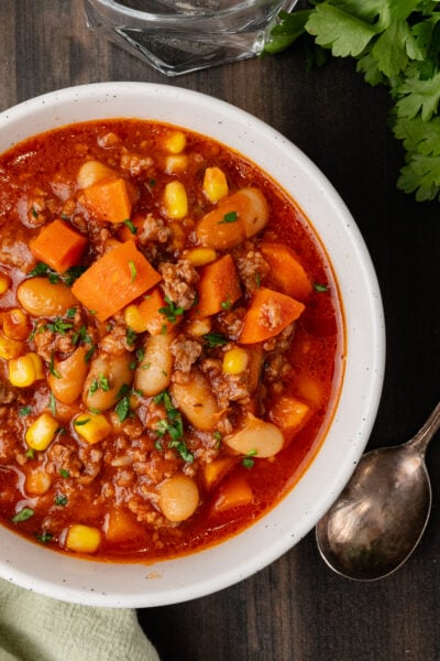 White bowl with ground beef, vegetables and bean soup on a wooden board with a silver spoon and fresh parsley.