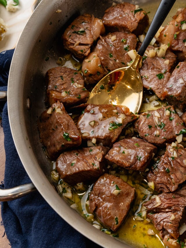 Metal saute pan with steak bites coated in butter, garlic and fresh parsley tossed with a gold spoon.