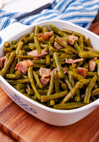 Southern style green beans and perfectly cooked chopped up bacon all sitting inside of a square serving dish sitting on a blue and white kitchen towel.
