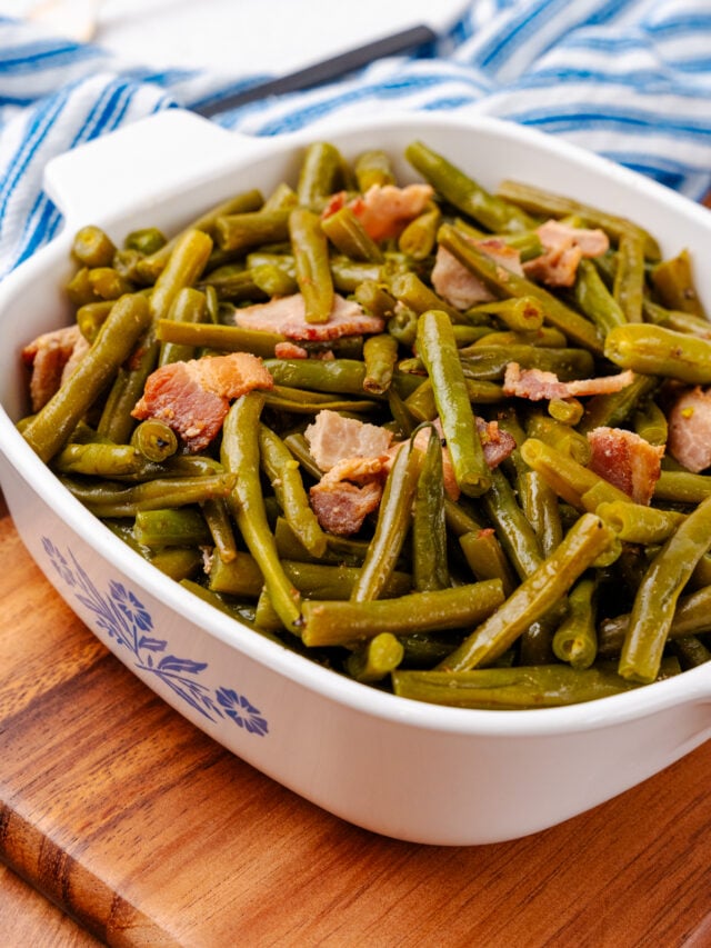 Southern style green beans and perfectly cooked chopped up bacon all sitting inside of a square serving dish sitting on a blue and white kitchen towel.