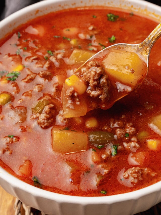 Vegetable beef soup in red tomato broth in a white bowl resting on a woven placemat.