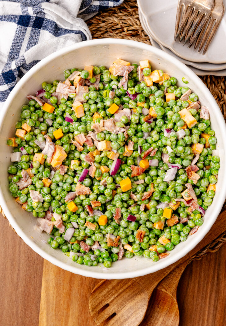 English pea salad 7 in white bowl with bacon, cheese, and peppers on wooden board with plates and napkin