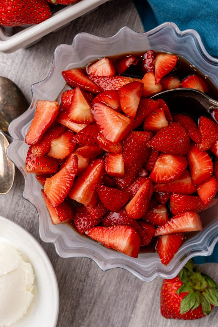 Balsamic strawberries 5 in a scalloped gray bowl with dark syrup, fresh berries and whipped cream nearby on gray surface