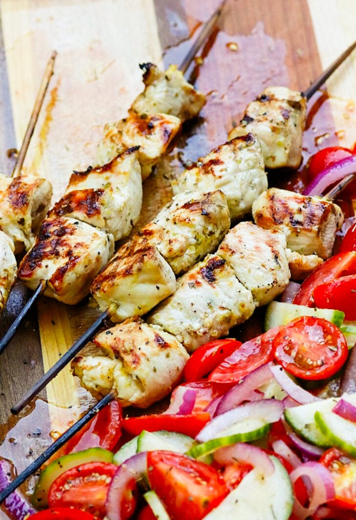 Greek chicken skewers 5 with char marks on wooden board, fresh tomato and cucumber slices alongside