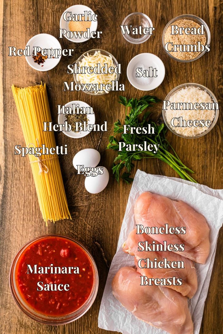 Overhead flat-lay of labeled ingredients for Crock Pot Chicken Parmesan — boneless skinless chicken breasts, marinara sauce, dry spaghetti, shredded mozzarella, grated parmesan, Italian bread crumbs, eggs, fresh parsley, Italian herb blend, garlic powder, red pepper flakes and salt arranged on a dark wood surface