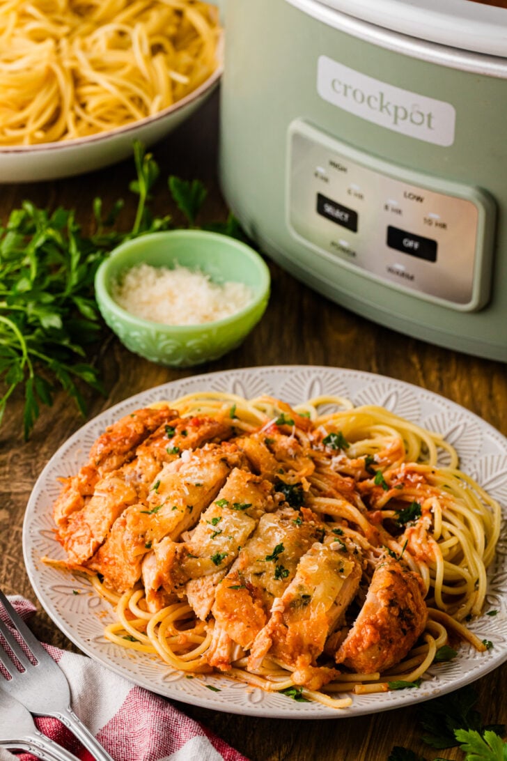 Plate of sliced Crock Pot Chicken Parmesan served over saucy spaghetti with grated parmesan and fresh parsley, a sage-green Crockpot slow cooker and a bowl of cooked spaghetti in the background on a rustic wood table with a red-striped napkin
