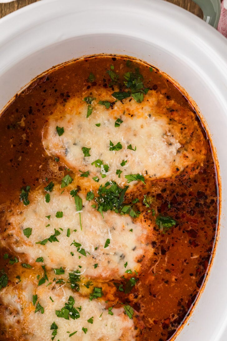 Overhead view of Crock Pot Chicken Parmesan — a golden breaded chicken breast smothered in melted mozzarella resting on a bed of marinara-tossed spaghetti, garnished with fresh chopped parsley and grated parmesan cheese on a rustic patterned plate