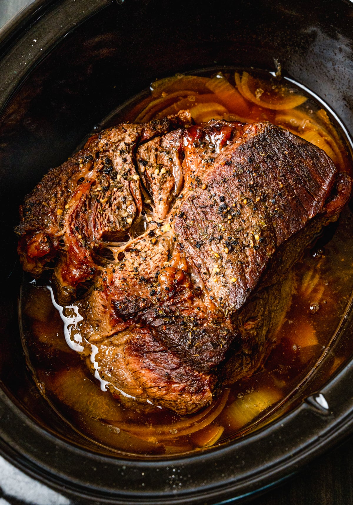 Slow cooker beef roast with caramelized onions and rich brown gravy in a black crockpot, overhead view