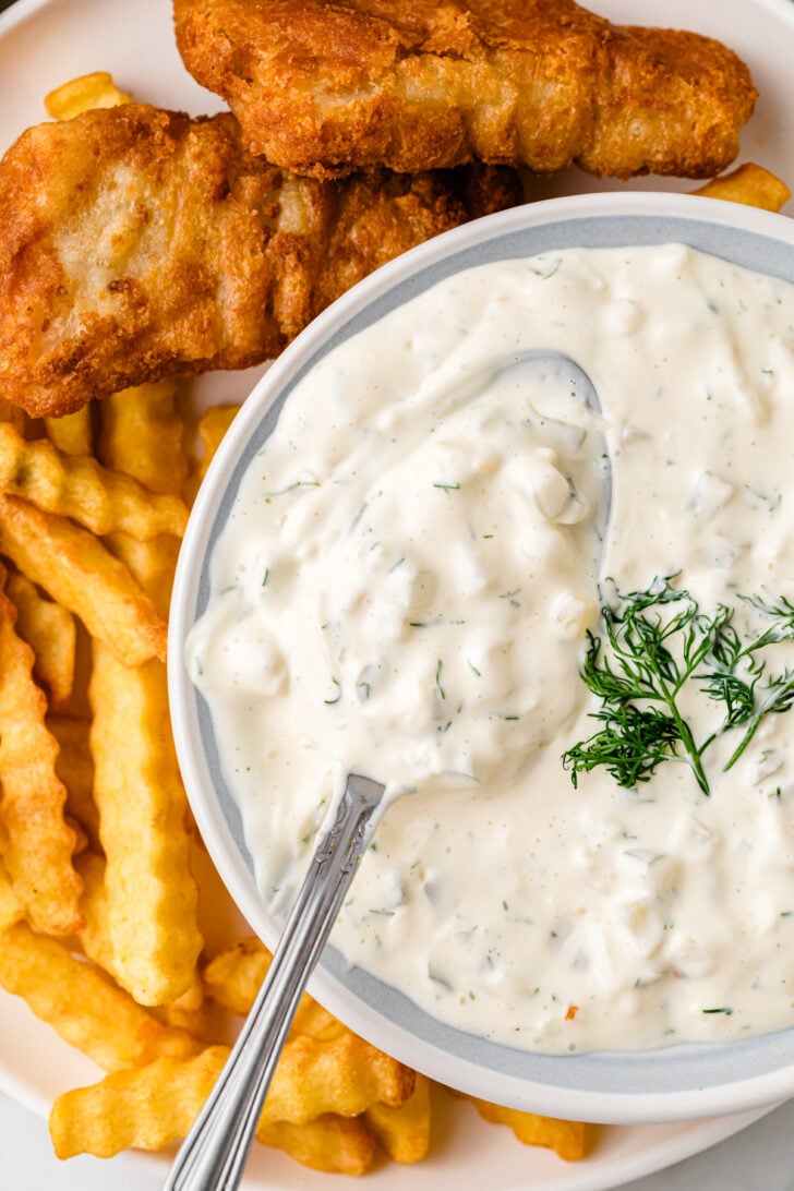 Easy Homemade Tartar Sauce in white bowl with spoon, crispy fried fish and golden fries on white plate