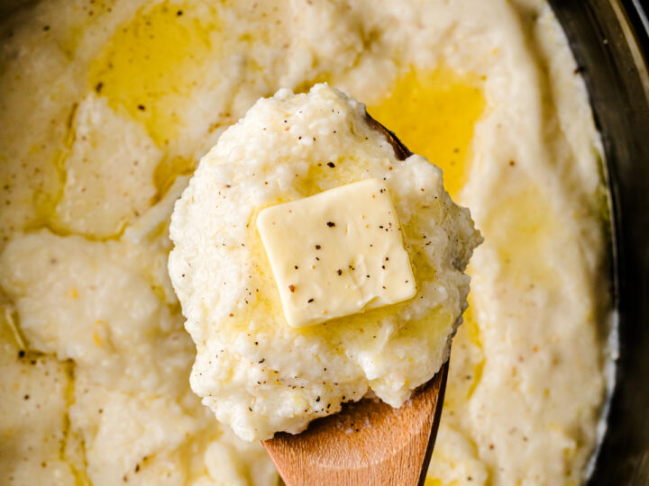 Crock Pot Southern Style Grits in black slow cooker with wooden spoon, topped with butter pat and black pepper