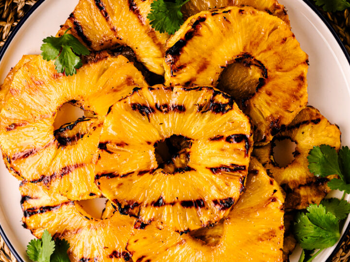 Grilled Pineapple with Brown Sugar Butter Glaze slices on white plate with cilantro garnish and cinnamon sticks on woven placemat